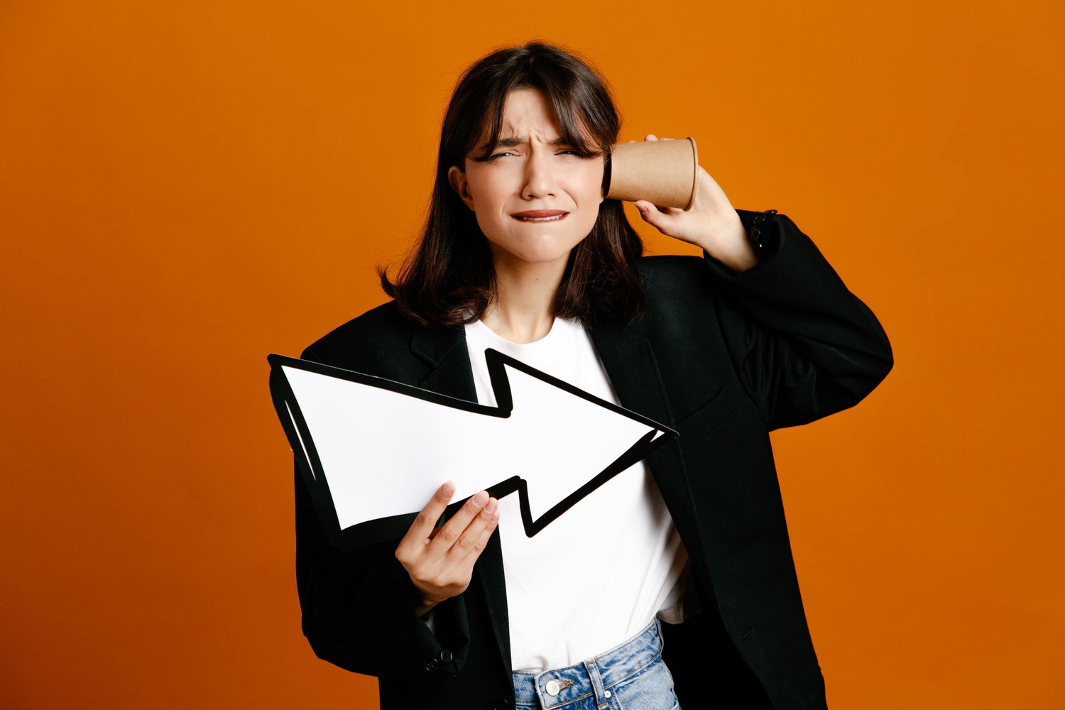 tense-putting-coffee-cup-ear-holding-direction-mark-young-beautiful-female-wearing-black-jacket-isolated-orange-background