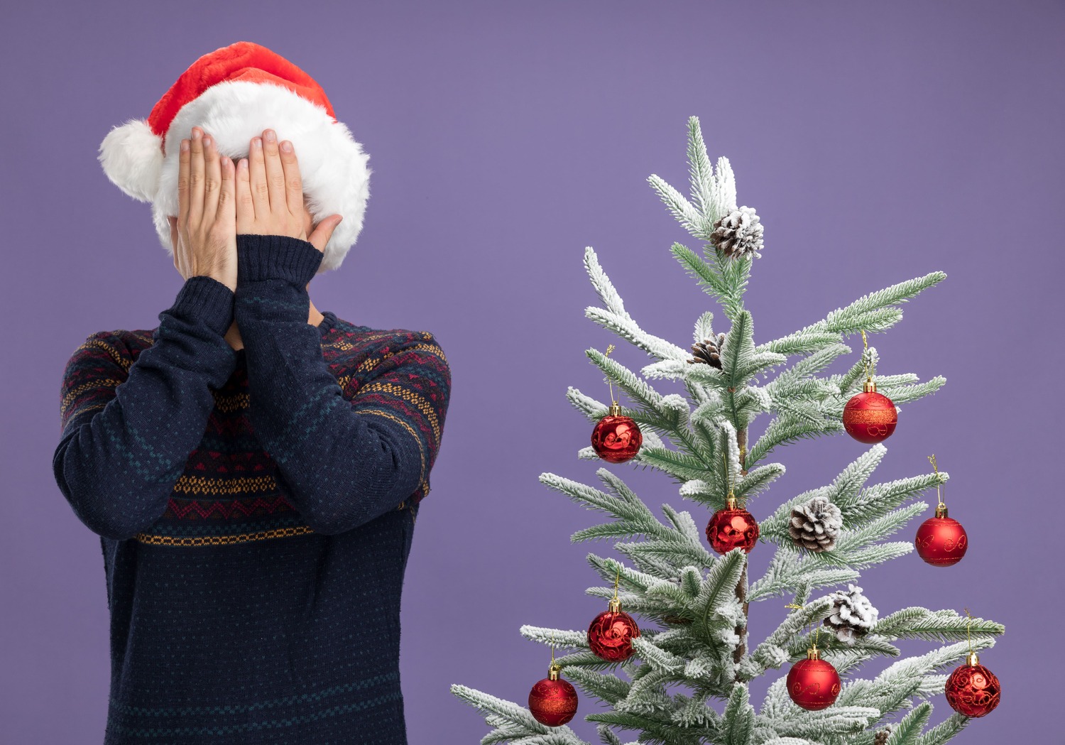 young-caucasian-man-wearing-christmas-hat-standing-near-decorated-christmas-tree-covering-face-with-hands-isolated-purple-background